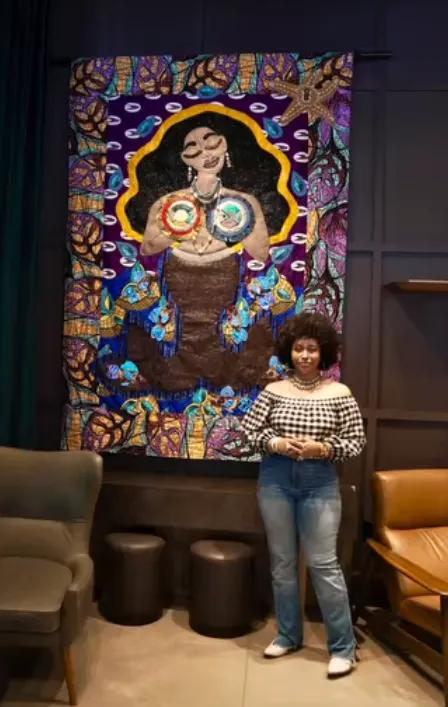 CC Mercer Watson standing beside her large beaded and stitched quilt PELA'S BREW, a portrait of a woman in purple, gold and blue